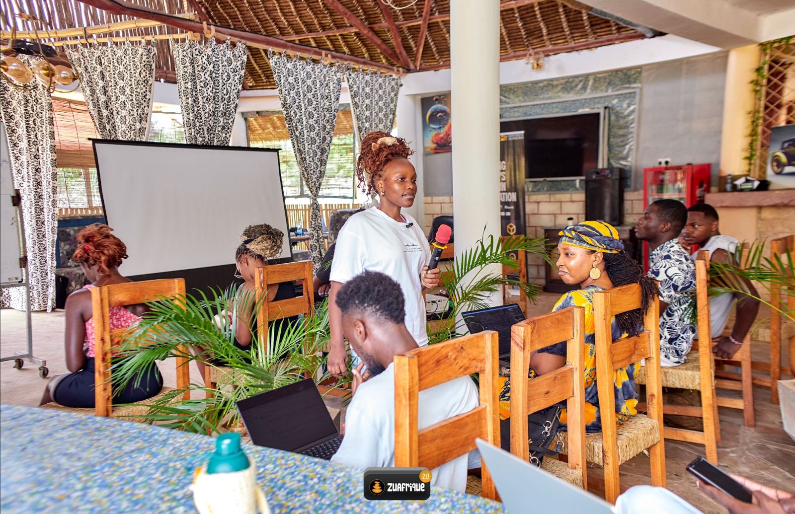 A resident leads a session in an open-air hall while others listen and take notes on laptops.
